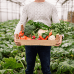 Homem em pé segurando uma caixa de madeira cheia de vegetais frescos e coloridos, como tomates, brócolis e cenouras, simbolizando a colheita saudável de um jardim orgânico livre de agrotóxicos.