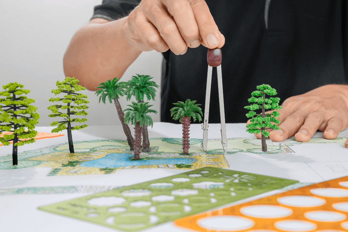 O paisagismo trabalha com a visão de futuro do jardim Mãos de um profissional utilizando compasso e miniaturas de árvores sobre uma planta baixa colorida, ilustrando como o paisagismo projeta a escala do tempo e o crescimento futuro da vegetação.